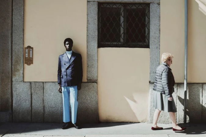 A man in a navy double-breasted blazer and light blue trousers stands against a wall as a woman walks past in profile.