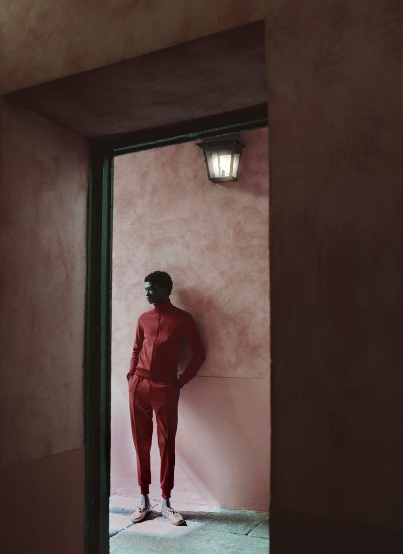 Model wearing a red tracksuit leaning against a textured pink wall framed by a dark doorway.