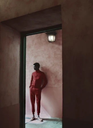 Model wearing a red tracksuit leaning against a textured pink wall framed by a dark doorway.