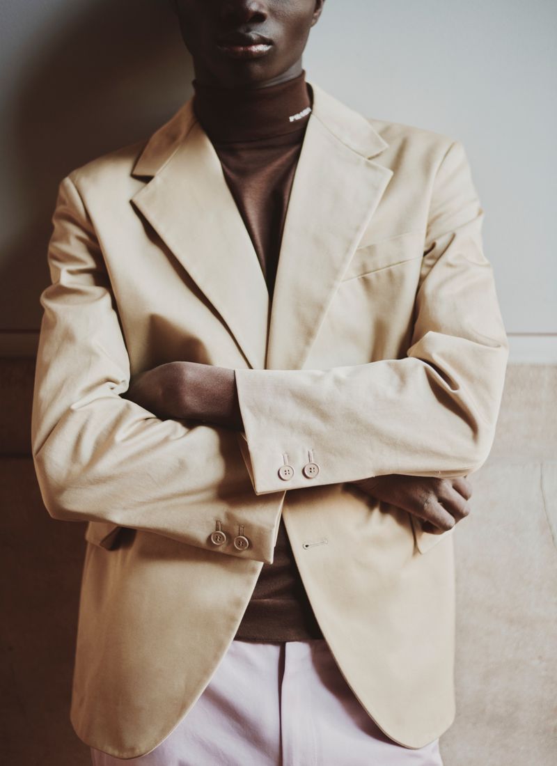 Man wearing a beige blazer and brown turtleneck with arms crossed against a plain interior background