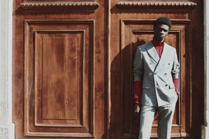 Model wearing a grey double breasted suit and red turtleneck standing before large wooden double doors.