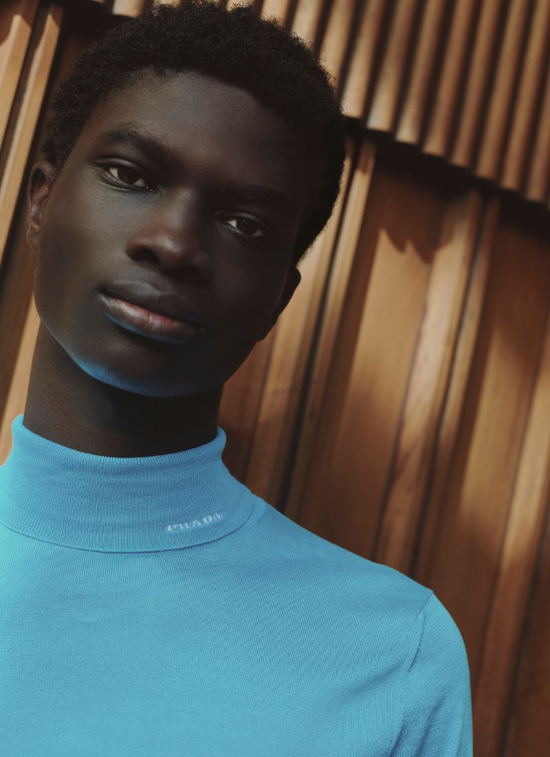 Close-up portrait of a Black man wearing a blue turtleneck shirt against a slatted wooden wall background.