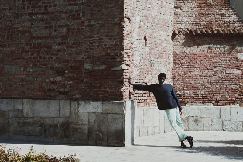 Model leaning against a weathered red brick building exterior wearing dark top and light trousers