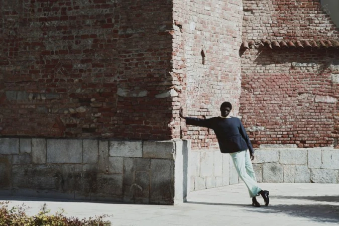 Model leaning against a weathered red brick building exterior wearing dark top and light trousers