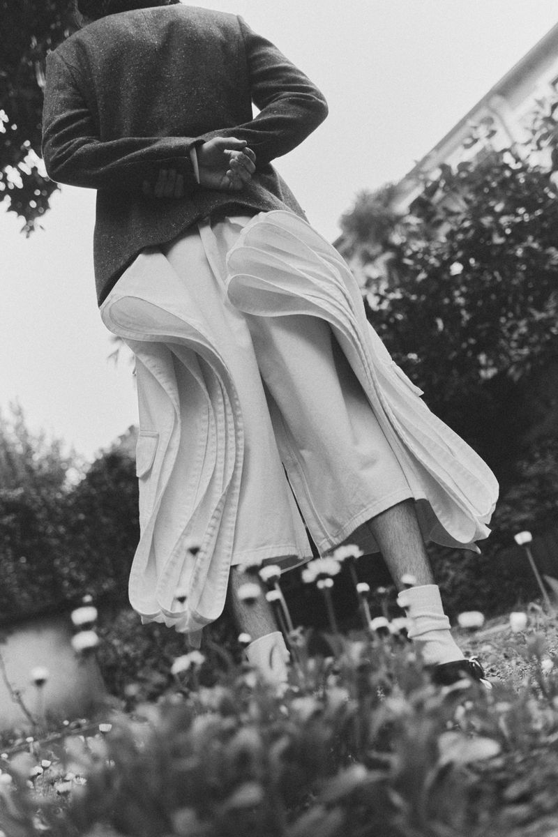 Model from a low angle wearing a dark blazer and flared white pleated trousers standing in an outdoor garden.