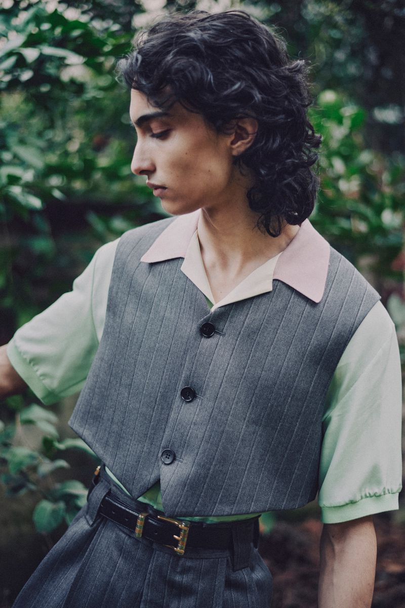 A young male model with dark wavy hair wearing a grey pinstripe vest over a pale green and pink collared shirt outdoors.