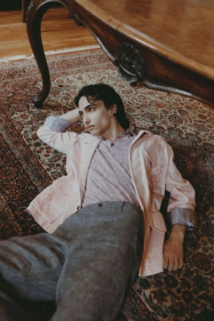 Male model lying on a patterned rug beneath a wooden table wearing a pink jacket and grey trousers