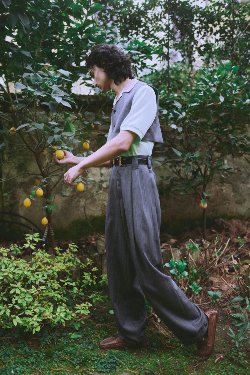 A young male model with dark curly hair wearing high-waisted grey trousers and a vest harvesting lemons from a tree in a lush garden.