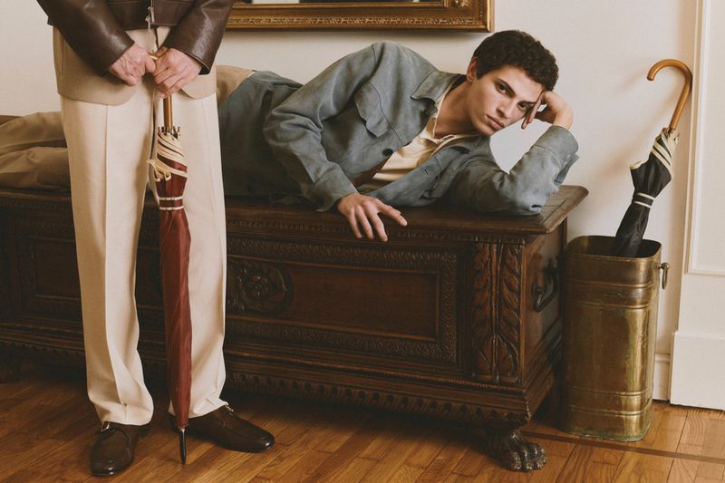 Two men posing indoors with umbrellas, one lying on a carved wooden chest wearing a grey jacket over a yellow polo.