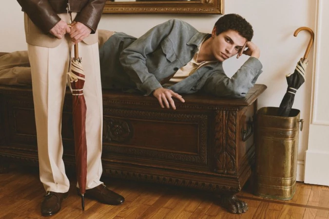 Two men posing indoors with umbrellas, one lying on a carved wooden chest wearing a grey jacket over a yellow polo.