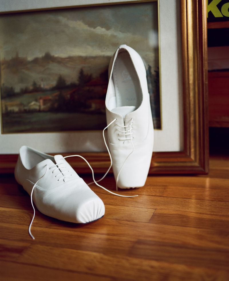 Two white leather dress shoes placed on a wooden floor in front of a framed landscape painting