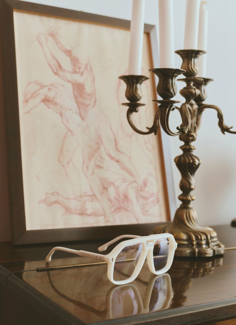 White framed glasses resting on a dark polished surface next to a brass candelabra and a framed drawing.