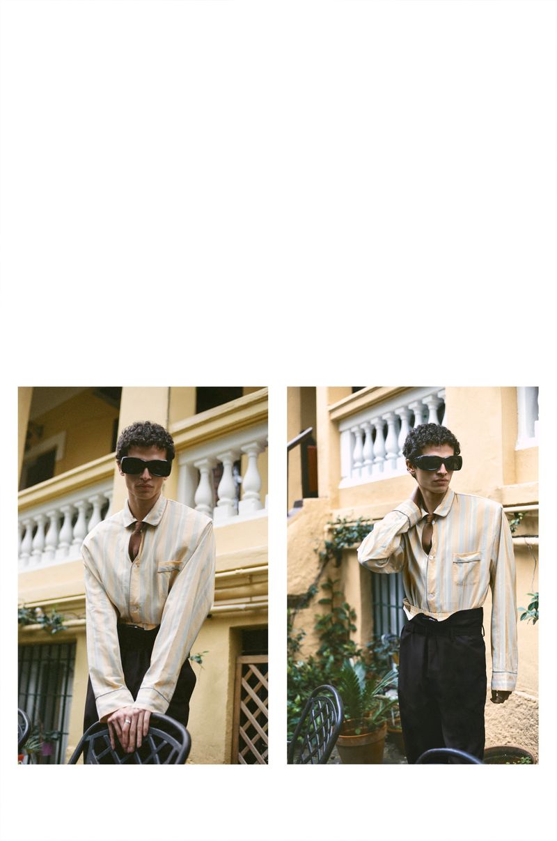 Model wearing dark sunglasses and a striped shirt leaning on a black chair against a cream building exterior.