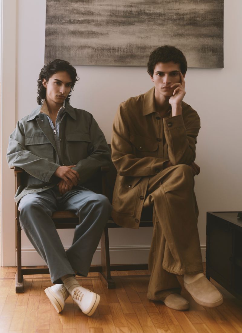 Two young male models seated on a dark wooden bench indoors, wearing menswear jackets and trousers against a white wall.