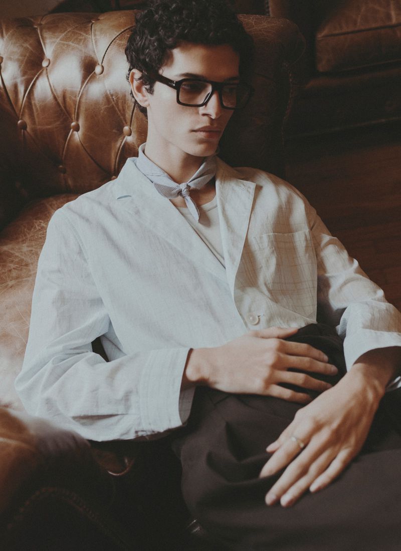 Man with curly hair and glasses in a white jacket lying on a brown leather couch indoors looking off to the side. Fashion photography.