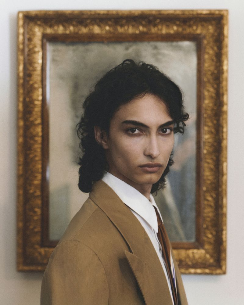 Portrait of a man wearing a brown suit jacket, white shirt, and tie with an ornate framed mirror behind him