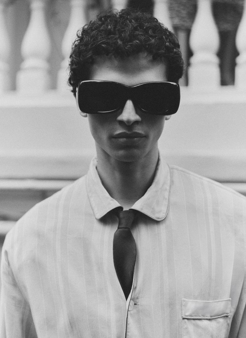 Portrait of a man wearing dark sunglasses, light collared shirt, and dark tie against an outdoor architectural balustrade