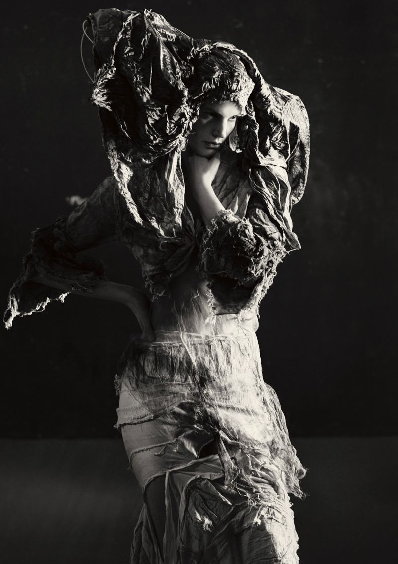 Monochrome fashion portrait of a woman in a draped fabric headpiece and textured dress, posing in a dark studio setting.