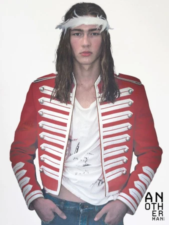 Long-haired male model wearing a red military-style jacket with silver buttons and a white feather headband against a plain background