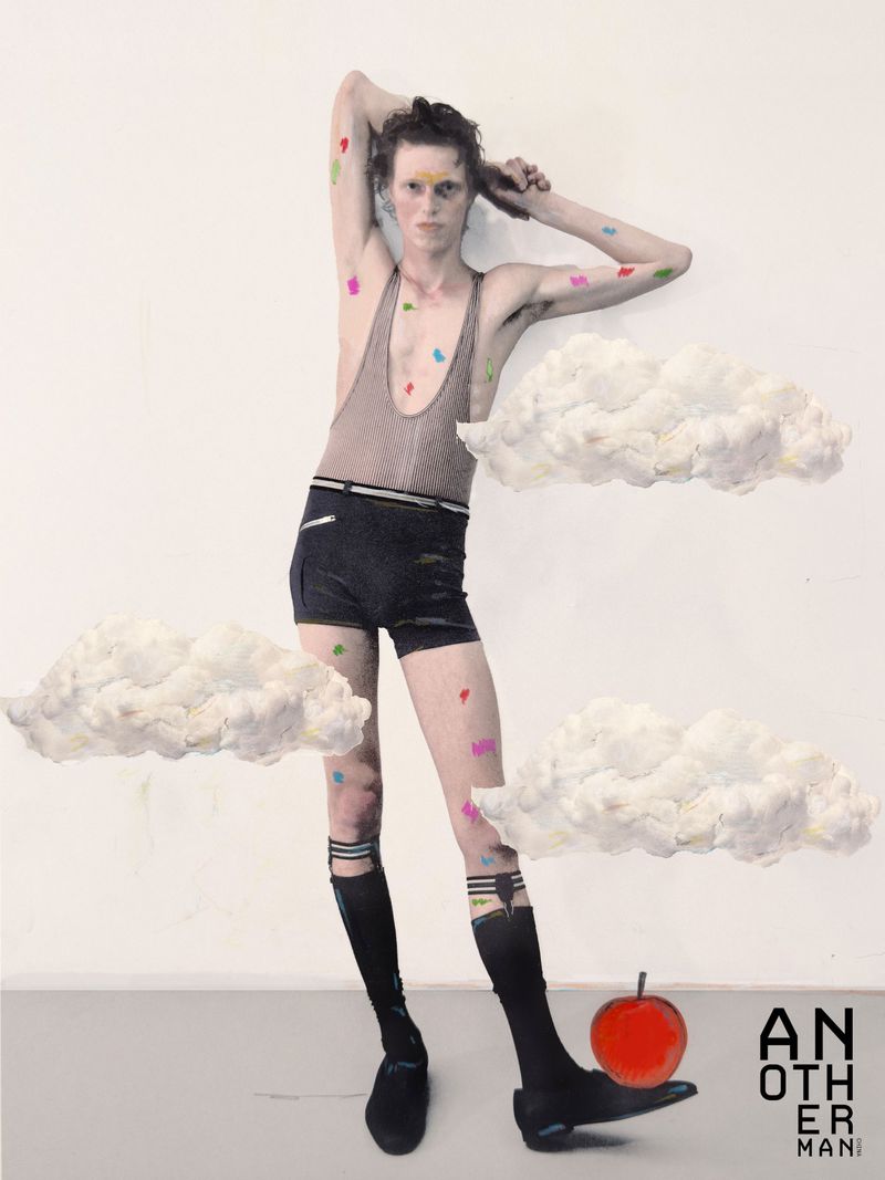 Male model in a taupe tank top and black shorts posing with paper cloud props against a white studio background