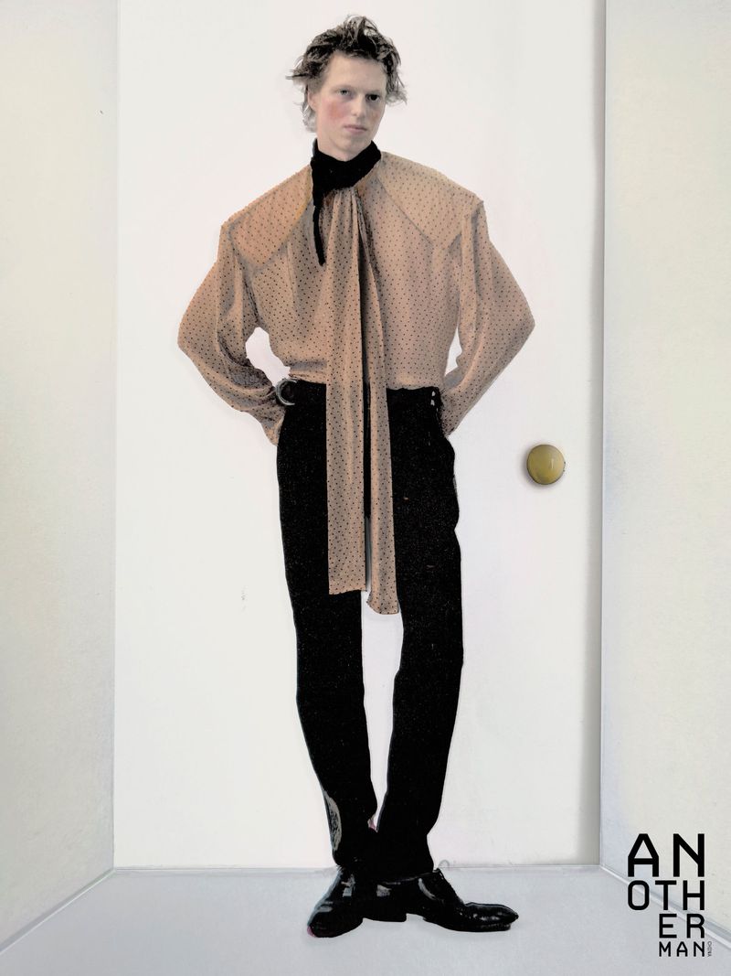 Model in beige polka dot blouse with long necktie and black trousers standing against a white door
