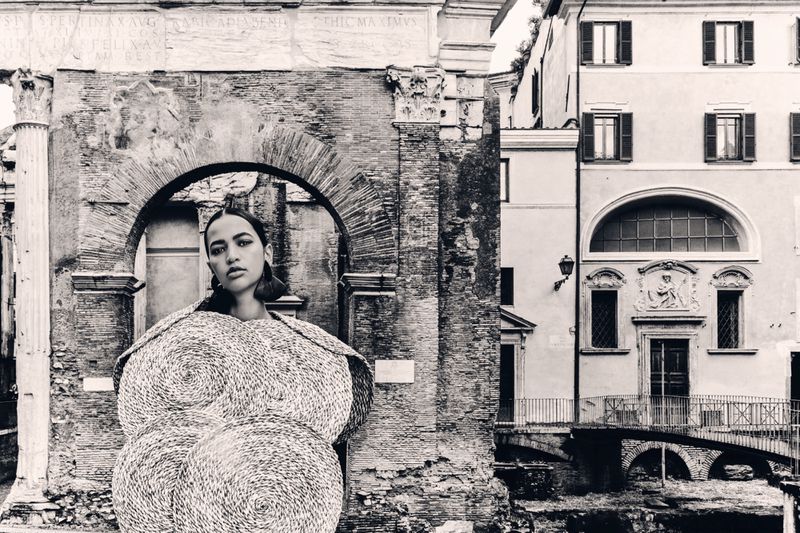 Model wearing sculptural textured circular garment framed by ancient stone archway and urban building