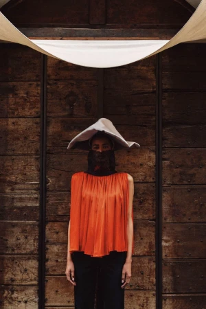 Model wearing a wide-brimmed hat, face veil, and orange pleated top against a dark wooden door for T Magazine MENA.