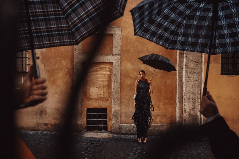 Model holding an umbrella standing against an ochre wall framed by other umbrellas in the foreground