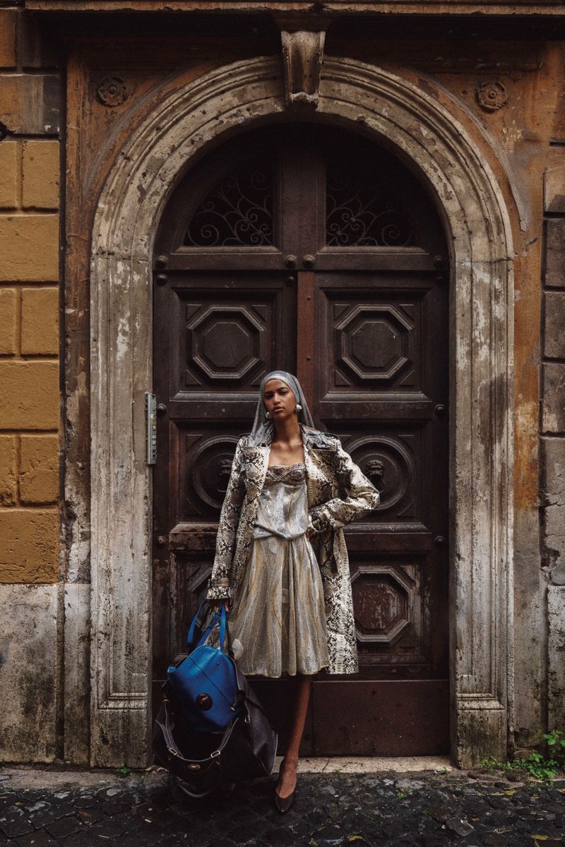 Model standing in front of an ornate arched wooden doorway wearing a patterned coat, holding a blue handbag.