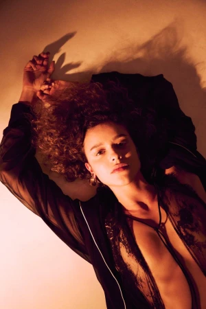 Overhead portrait of a curly-haired woman lying down in dark clothing with warm directional lighting casting shadows against a pale surface.