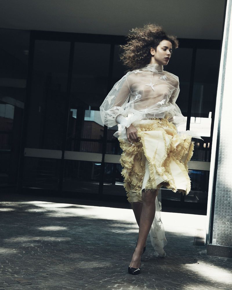 Model with voluminous curly hair wearing a sheer white patterned blouse and a ruffled yellow skirt walking outside