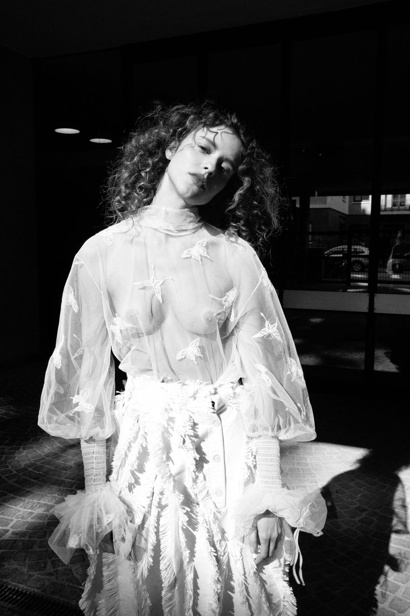 Black and white portrait of a woman with curly hair wearing a sheer patterned blouse and light skirt.