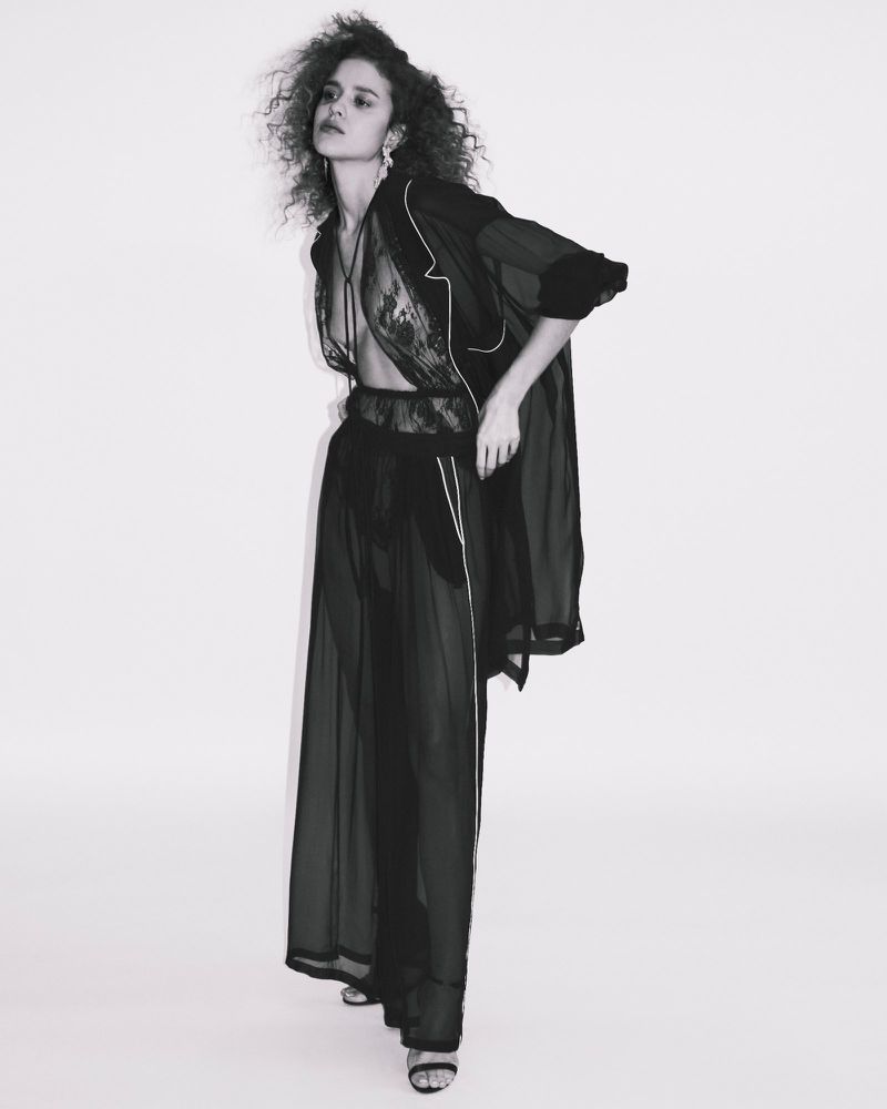 Model with curly hair standing in sheer black lace trousers and matching robe against a clean white background.