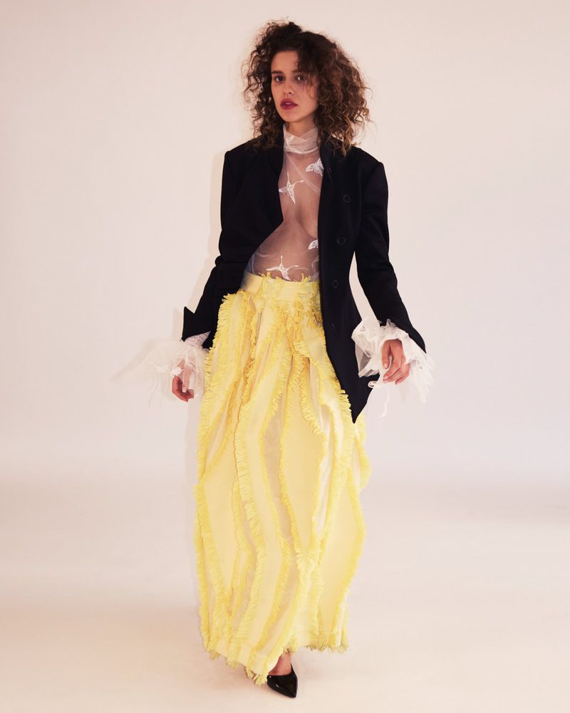 Model with curly brown hair wearing a black blazer and bright yellow pleated skirt against a white studio background.