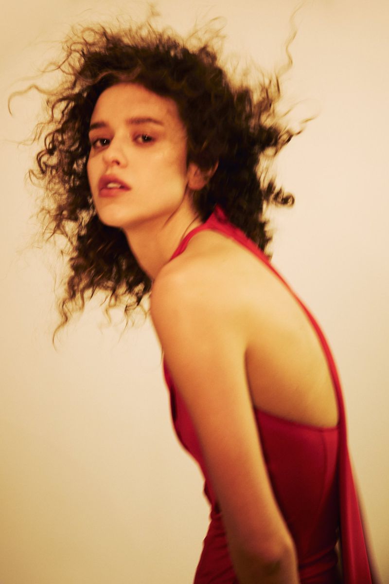 Woman with curly dark hair wearing a bright pink sleeveless dress against a plain neutral background.