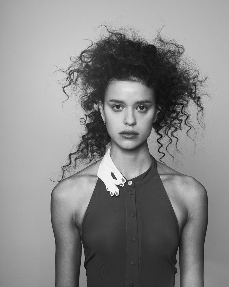Portrait of a young woman with curly hair wearing a red dress with a white collar against a gray background in black and white.