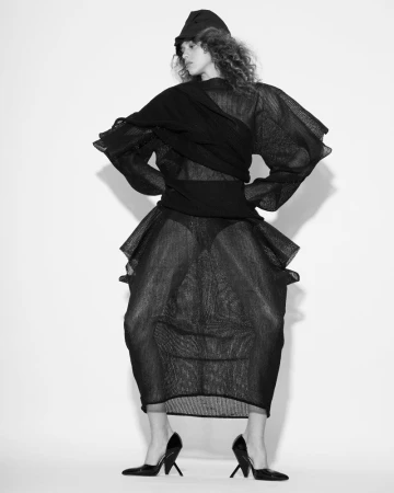 Model in sheer black structured dress, cap, and heels against a white seamless background for Elle Slovenia