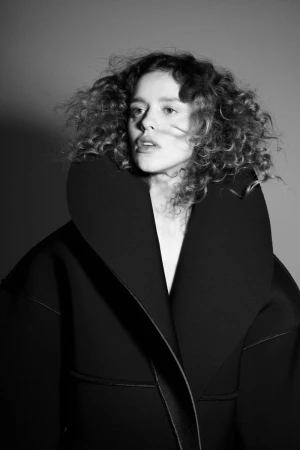 Monochrome portrait of a woman with curly hair wearing a structured high-collar black coat against a grey studio backdrop.