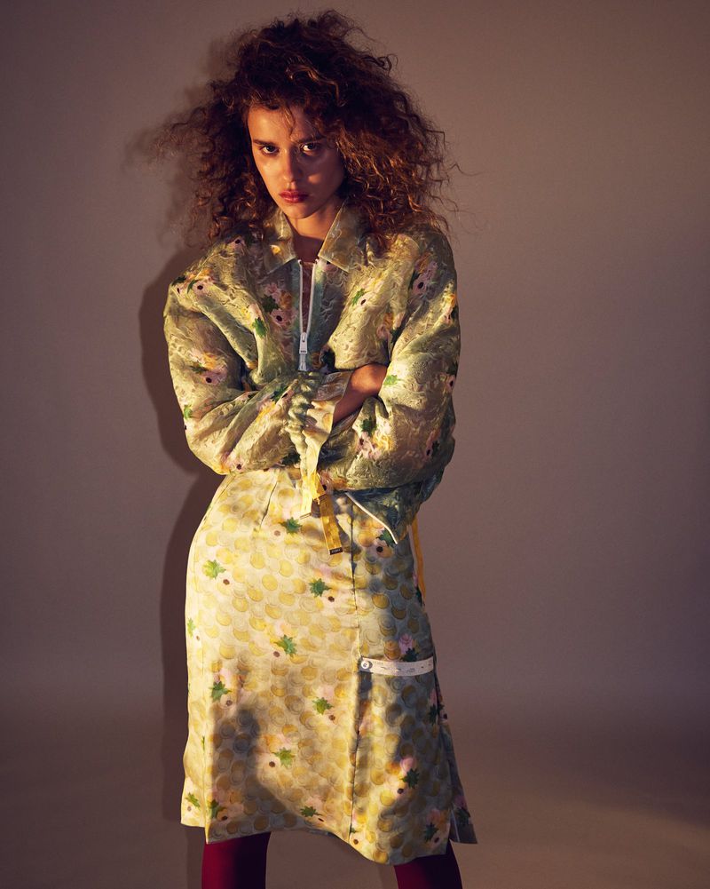 Woman with curly hair wearing a patterned green jacket and matching skirt against a plain studio background.