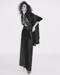 Model with curly hair standing in sheer black lace trousers and matching robe against a clean white background.