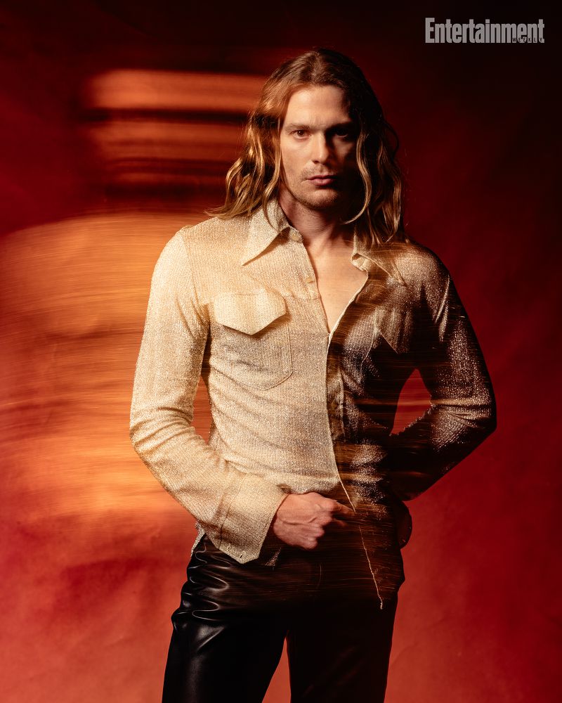 A long-haired man with a serious expression wearing a metallic gold shirt and black pants against a red backdrop