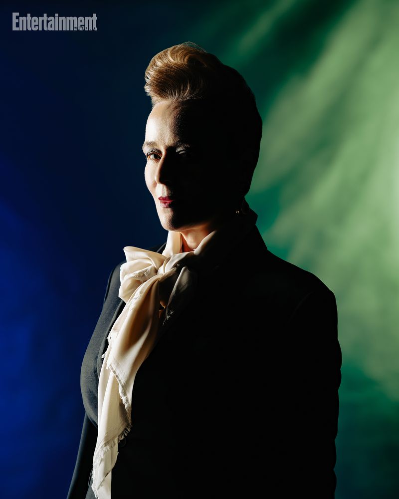 Portrait of a person wearing a dark suit and ascot against a blue and green lit background for Entertainment Weekly.