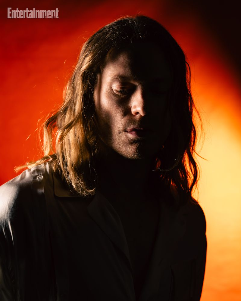 Portrait of a man with long hair in a dark shirt against a warm red and orange gradient background for Entertainment Weekly.