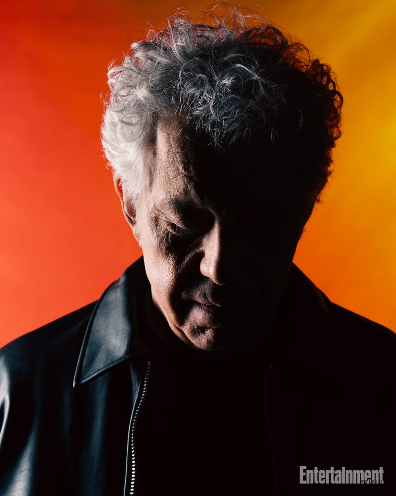 Portrait of an older man with curly hair wearing a black leather jacket against an orange and yellow gradient background.