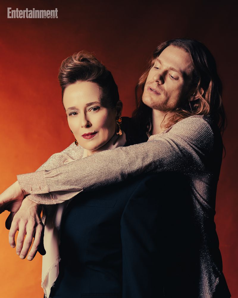 Two people posing against a warm gradient background for Entertainment Weekly, featuring a woman seated and a man embracing her from behind.