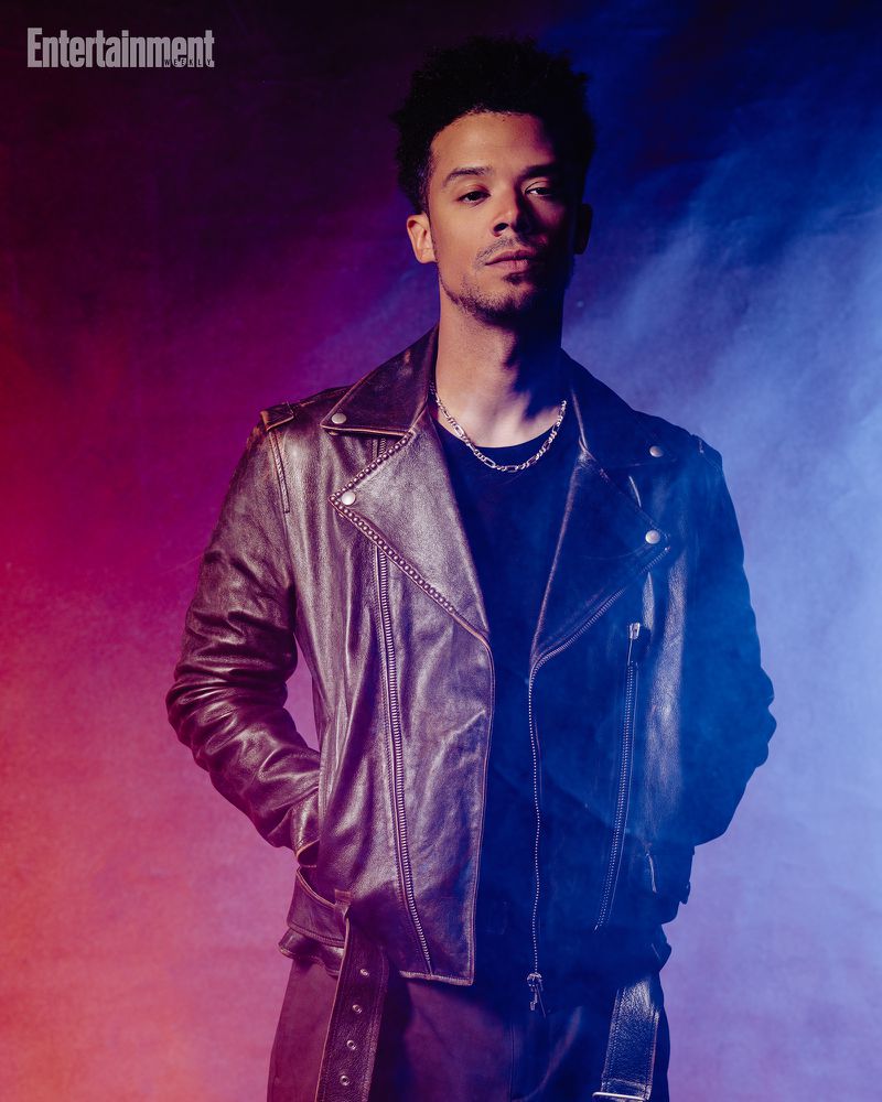 Man wearing a brown leather jacket and silver chain necklace against a moody purple and blue background.