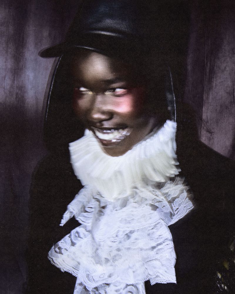 Portrait of a black woman wearing a white lace ruffled collar and black cap with dramatic flush makeup.