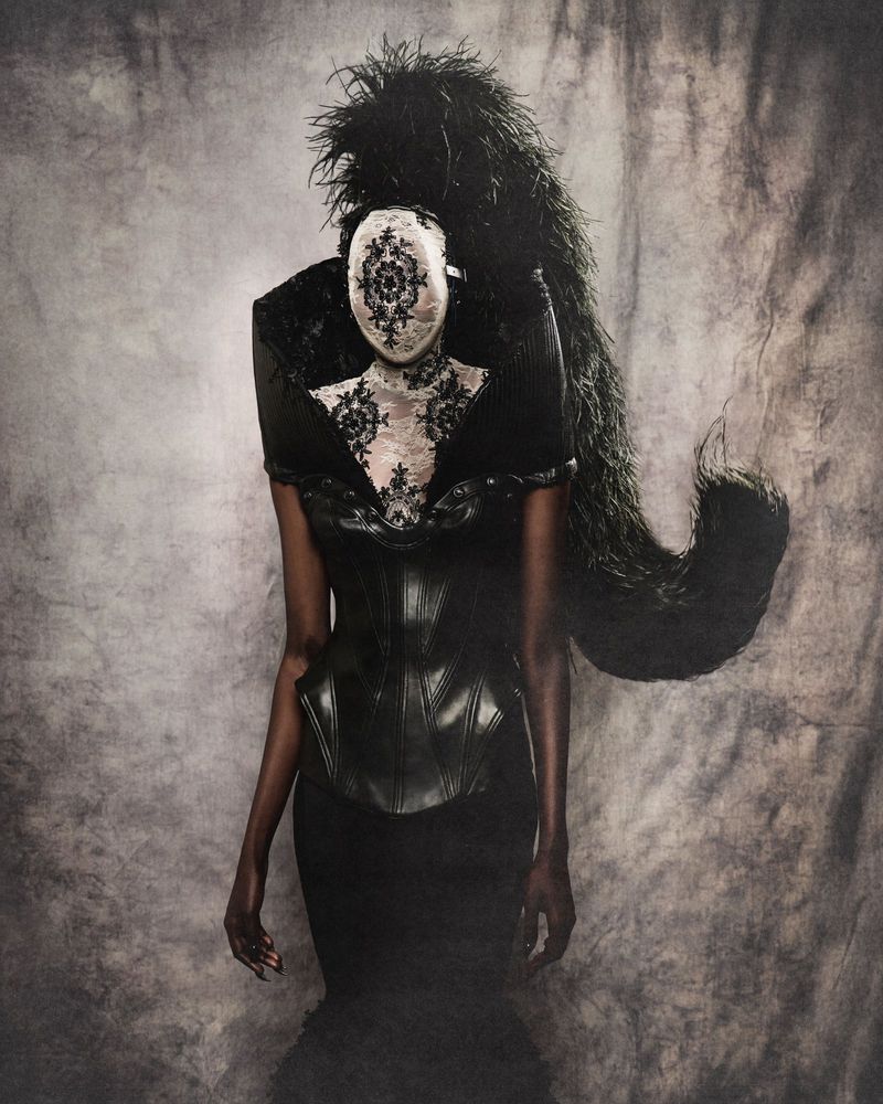 Person in black corset dress with ornate lace mask and large feathered headdress against a textured gray backdrop in a fashion portrait.