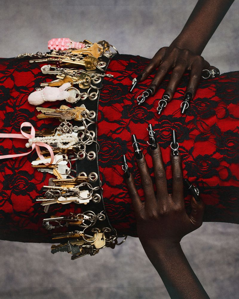 Close-up of a person with black nail rings resting hands on a red lace dress with a key-covered belt for Swallow The Key