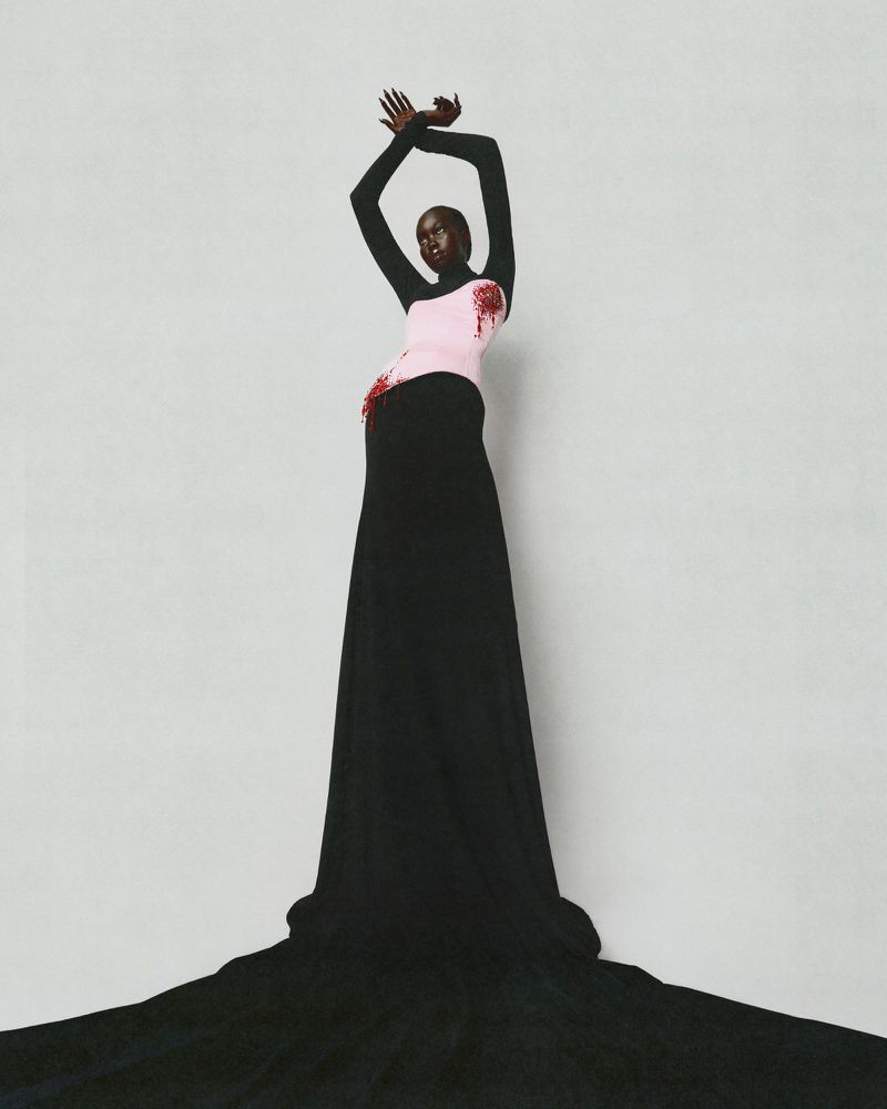 Model in a black long-sleeve top and pink corset with black floor-length skirt against a grey studio backdrop.